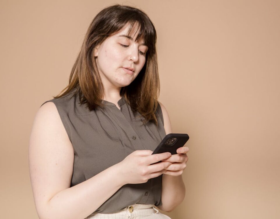 Focused plump Asian female in stylish wear text messaging on modern cellphone while sitting on beige background in light studio