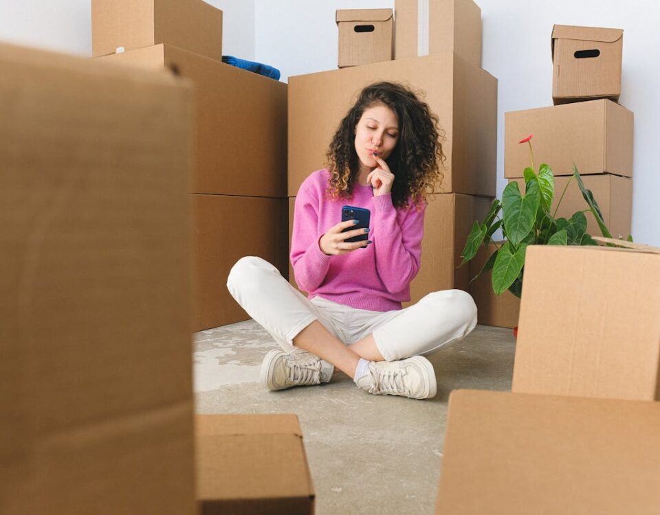 5 apps para encontrar o imóvel dos seus sonhos Full body of positive thoughtful young female millennial in casual outfit touching lips and using smartphone while sitting on floor with crossed legs near carton boxes after relocation