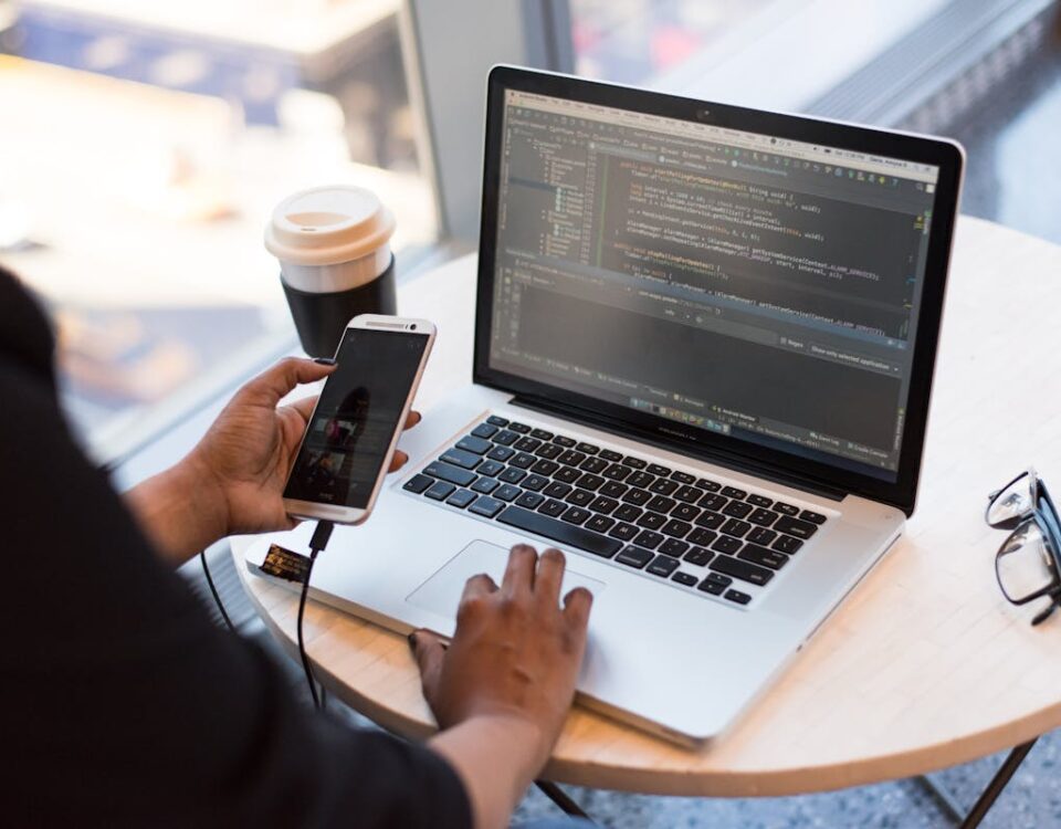 5 softwares de backup em nuvem que você precisa conhecer Developer working remotely, coding on a laptop with phone in hand, showcasing modern work culture.