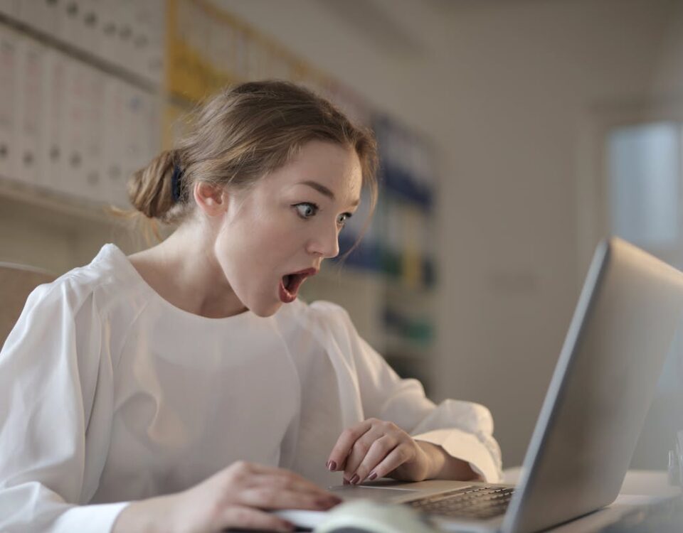 Qual a importância de um detetive virtual? Surprised woman sitting at desk with laptop indoors, expressing amazement.