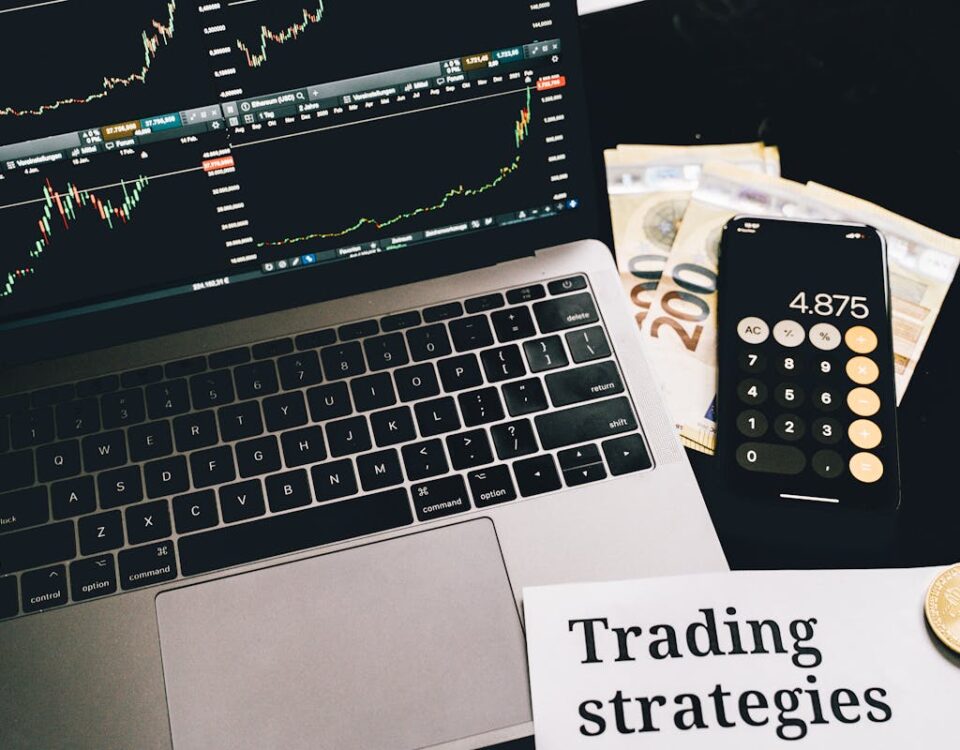 5 apps para economizar e investir dinheiro A laptop showing stock market graphs with a smartphone calculator, euro bills, coins, and trading strategies paper.