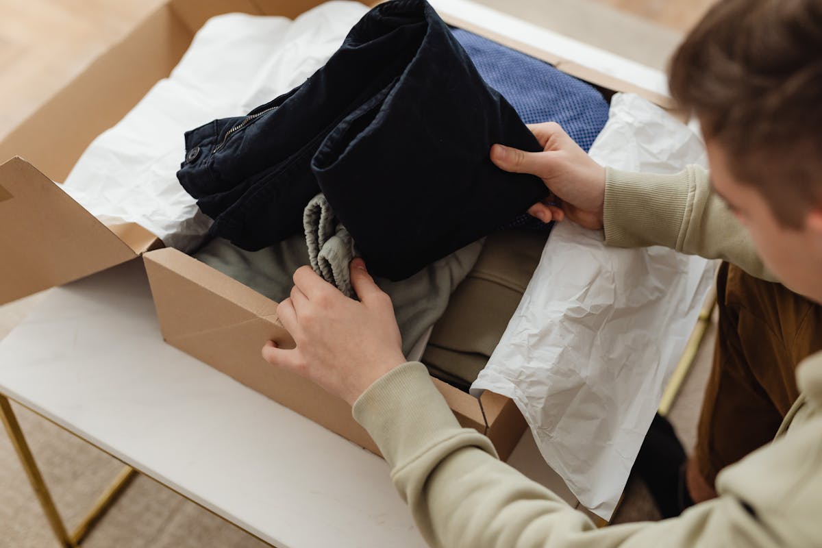 5 apps para comprar e revender roupas infantis Person unpacking clothes from a delivery box indoors, showcasing a range of garments.