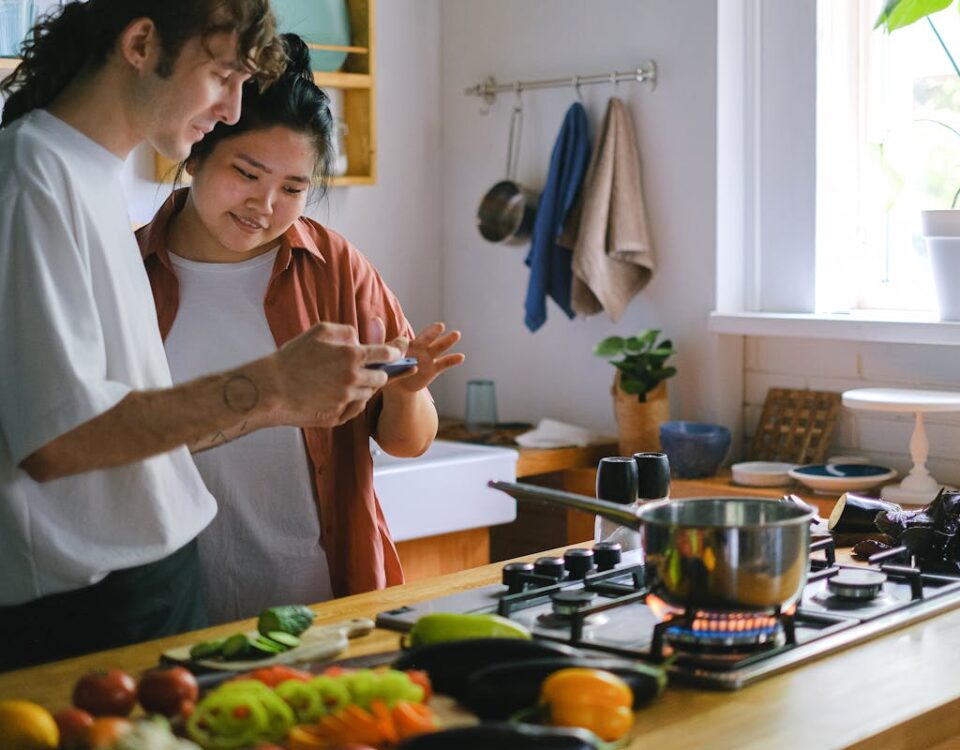 5 aplicativos que ajudam a organizar receitas e o dia na cozinha A young couple uses a phone while preparing a meal in their modern kitchen with fresh vegetables.