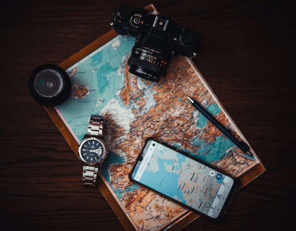 5 apps para criar lembranças personalizadas de viagens Top view of travel essentials including a map, camera, phone, watch, and lens on a wooden surface.