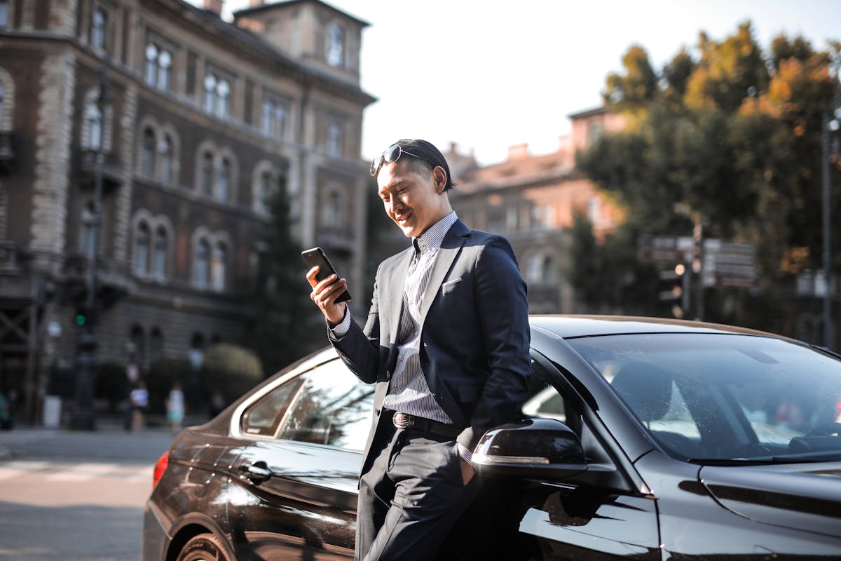 5 apps para gerenciar a validade da sua CNH Smiling businessman in a black suit texting on his phone while leaning against a parked car in the city.
