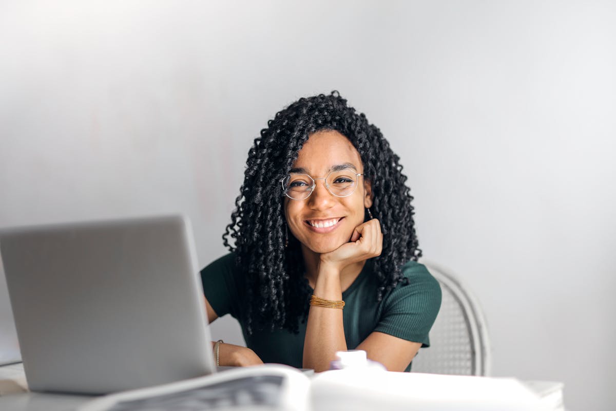 5 aplicativos para estudar para a prova da CNH Joyful businesswoman with curly hair smiling at camera while using laptop indoors.