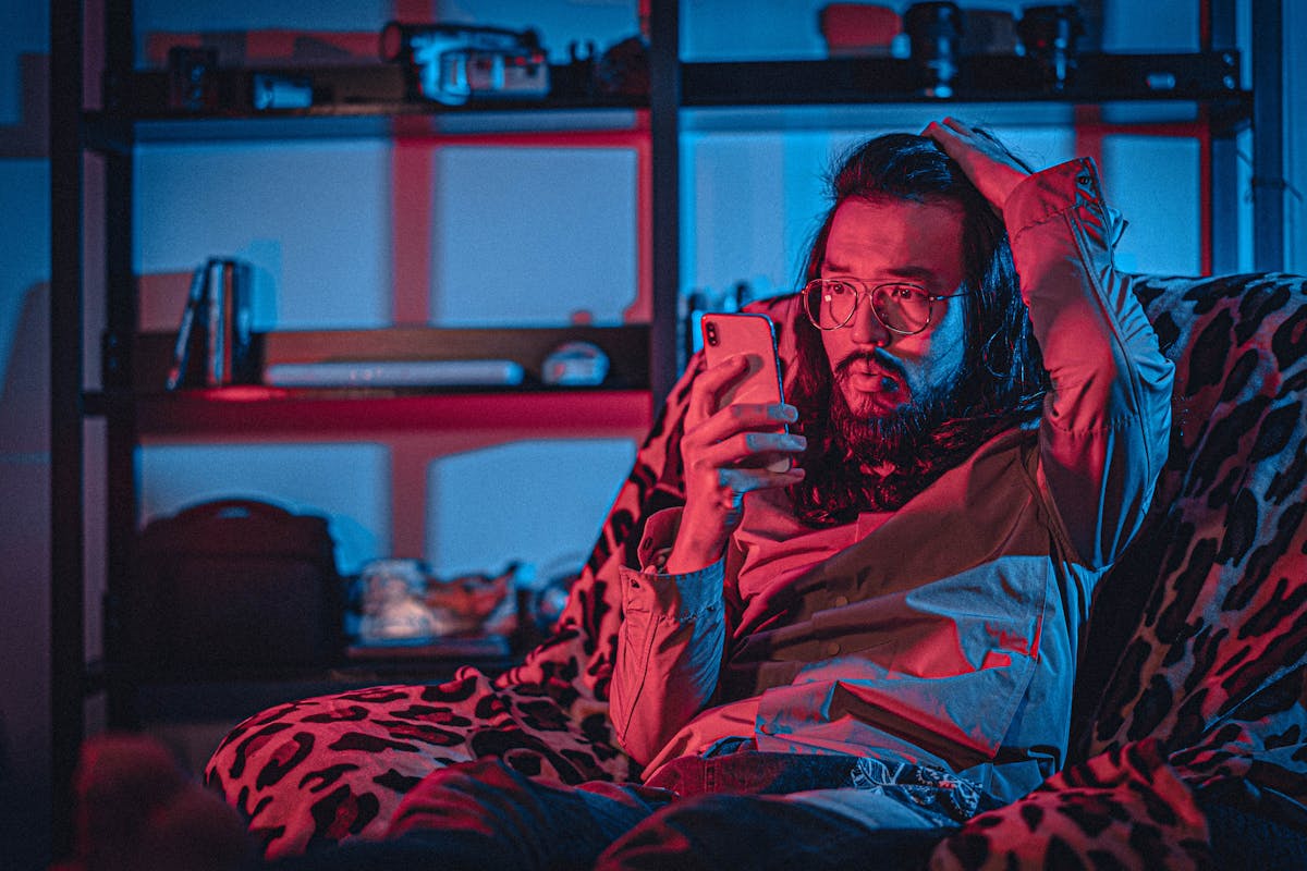 Surprised adult unshaven ethnic male in eyeglasses with pouting lips browsing internet on cellphone while touching head and sitting on comfortable armchair near shelf in apartment with neon light