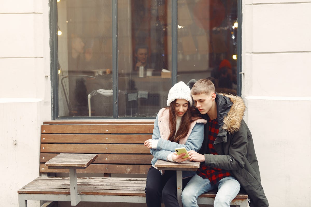 A young couple in warm clothing looks at a smartphone together on a bench outside a cafe.