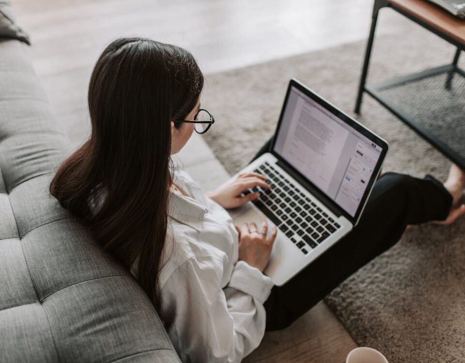 5 aplicativos para estudar para a prova da CNH Woman working remotely with a laptop on the floor next to a sofa, enjoying comfortable home office setup.