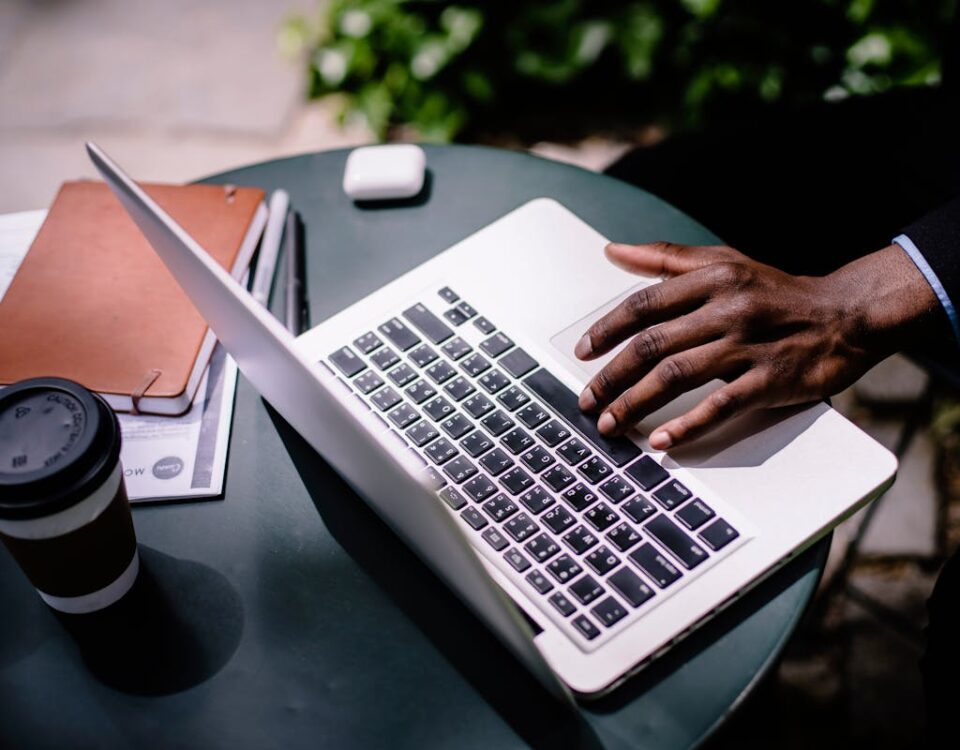 5 softwares que facilitam a gestão de seguros e planos A businessman uses a laptop at an outdoor cafe table with coffee and documents nearby.