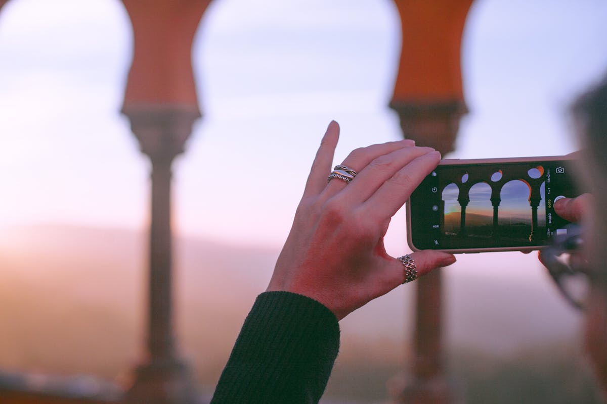 Crop unrecognizable female traveler taking photo of old oriental castle with arched passage on mobile phone against sunset sky