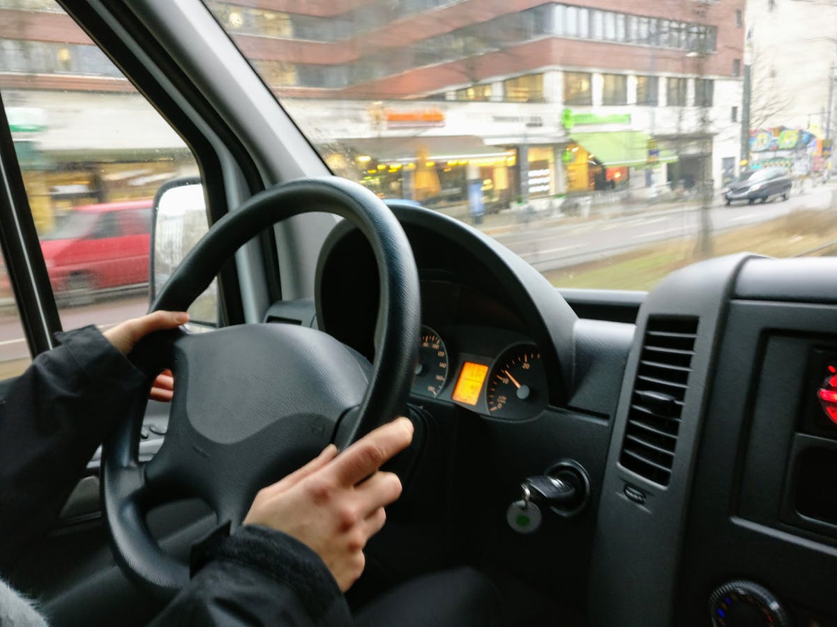 5 apps que ensinam técnicas de direção defensiva Hands on a steering wheel driving a van through an urban area with shops visible outside.