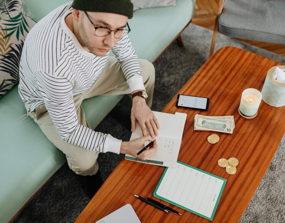 5 plataformas para acumular milhas e cashback em 2025 A man in a striped sweater sits on a sofa, writing in a notebook with cash and coins on a wooden table.