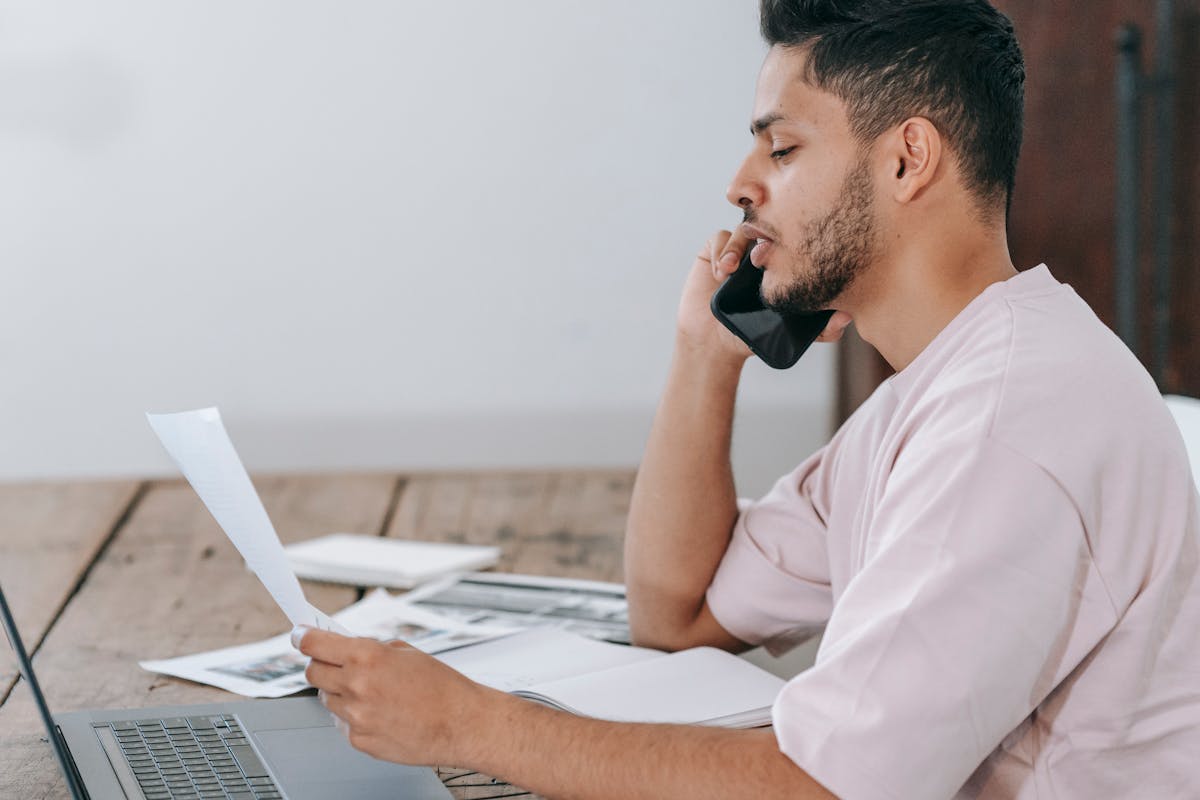 5 apps para comparar seguros e economizar Side view of young Hispanic male remote employee discussing documents during phone call while working online with laptop and papers in home office