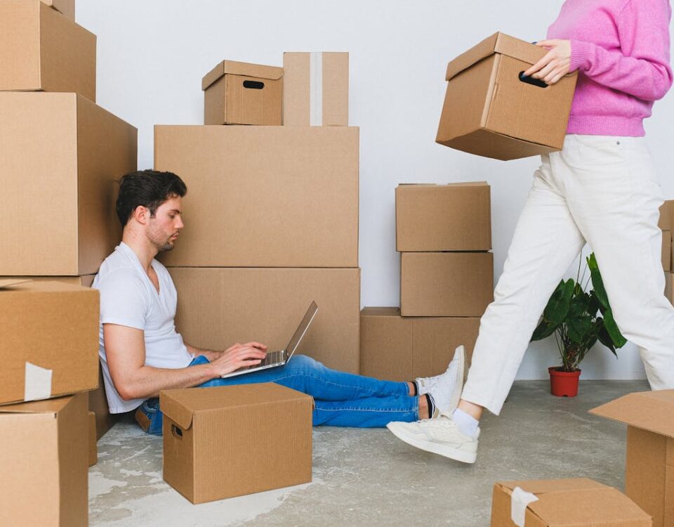 5 aplicativos para organizar a mudança de casa Young couple settling in new apartment surrounded by packing boxes, managing tasks on a laptop.