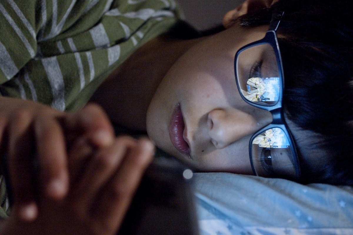 5 melhores apps para assistir filmes no celular A child lying in bed at night using a smartphone, with screen reflection on eyeglasses.