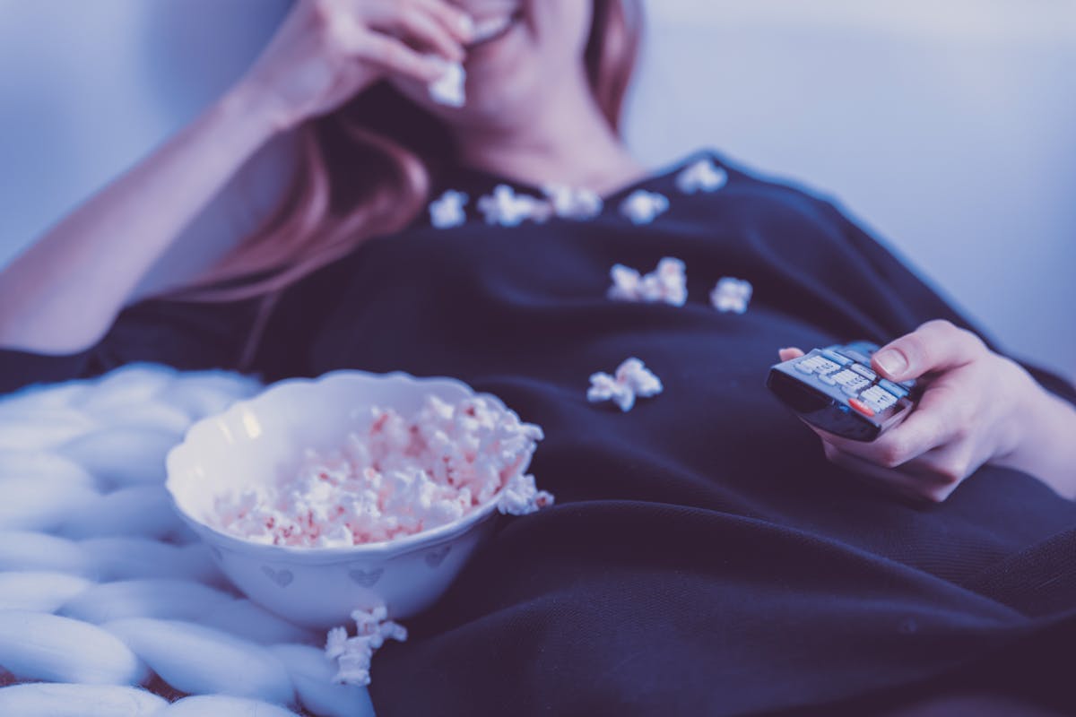 Woman relaxing on couch eating popcorn while watching TV, holding remote control, indoors.