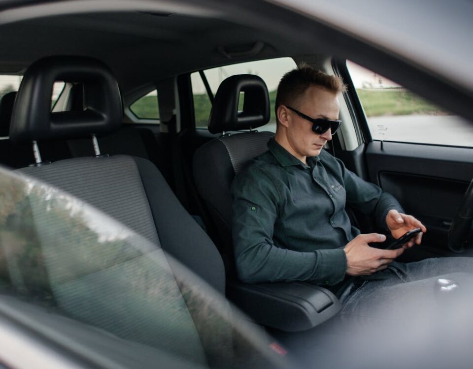 5 apps essenciais para quem está tirando a CNH Young man using smartphone inside a parked car, wearing sunglasses.