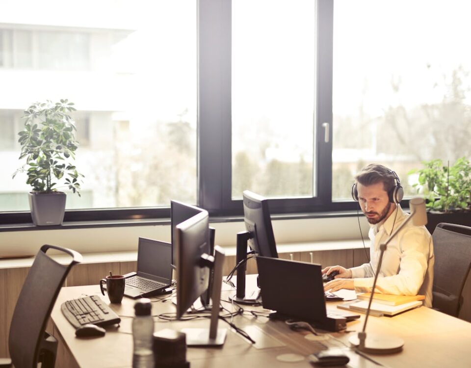 Softwares que organizam e potencializam sua central de atendimento A businessman sits at a desk using multiple computers and a headset in a well-lit modern office.