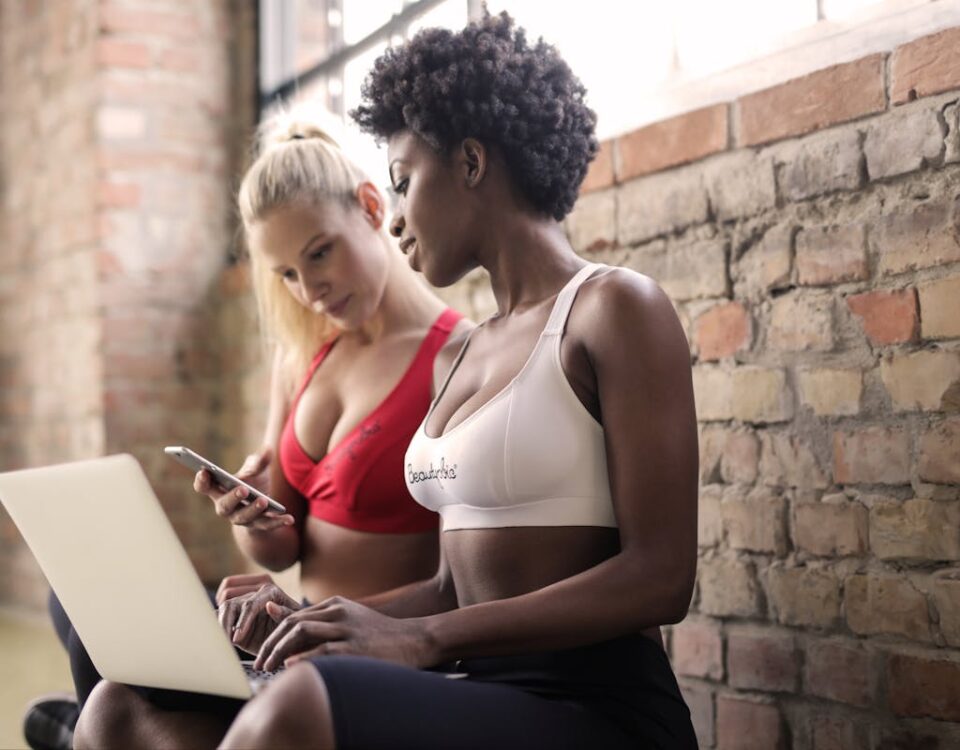 5 tipos de softwares para gestão de arenas esportivas Two women in sportswear using a laptop and smartphone against a brick wall, showcasing fitness and collaboration.