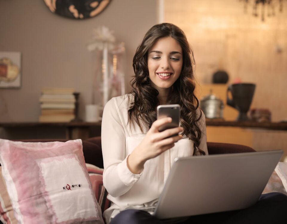 5 softwares para uma administração mais eficiente A woman enjoying leisure time using her smartphone and laptop in a cozy living room.