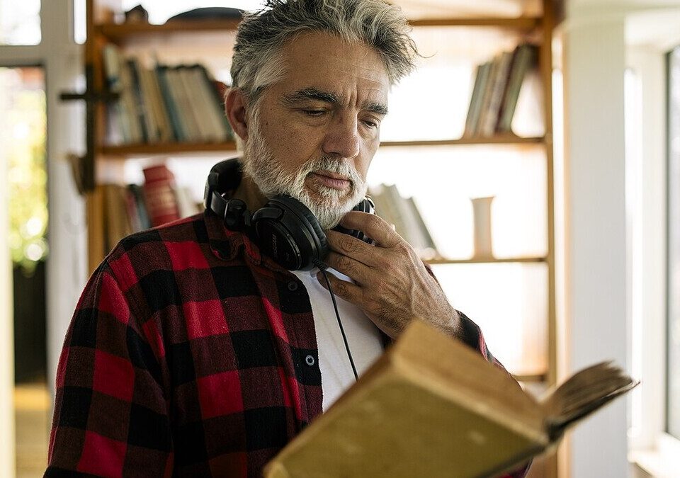 Audiolivros no Spotify chegam ao Brasil: como usar as 12 horas grátis Homem com fones de ouvido no pescoço lendo um livro em frente a uma estante