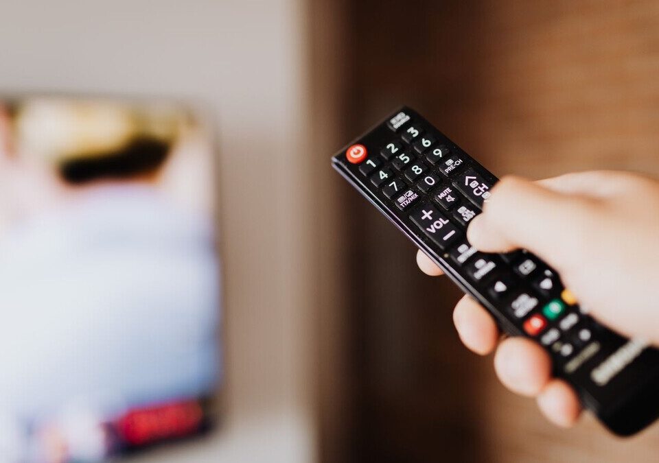 Mão apertando botão de controle remoto diante de uma TV