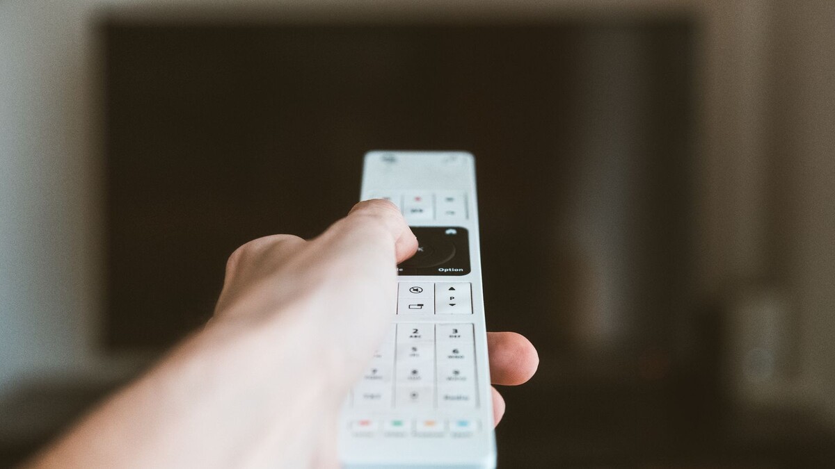 Mão segurando controle remoto diante de uma TV ligada